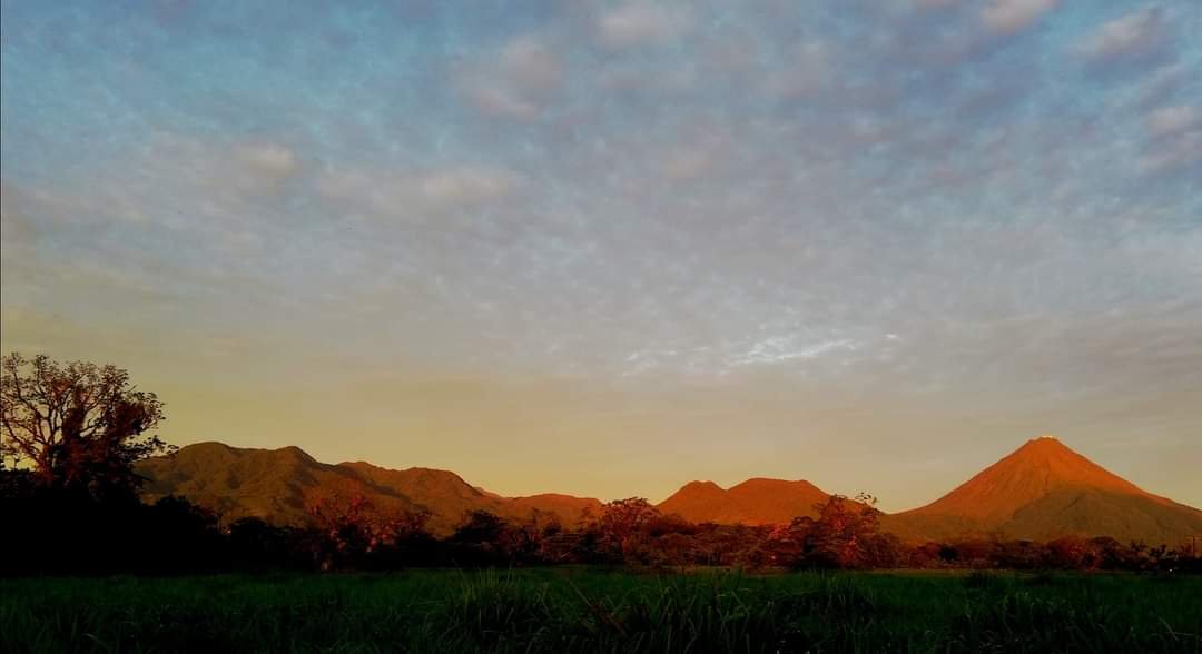 A scenic view of the Arenal mountain range during sunset, with the peaks bathed in warm golden light against a partly cloudy sky.